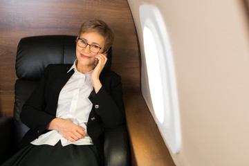 beautiful aged woman in the cabin of a private plane in an armchair with a phone in her hands