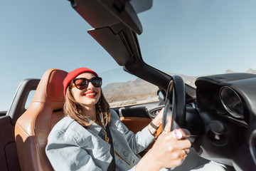 Happy woman in red hat driving convertible car while traveling on the desert road. Carefree...