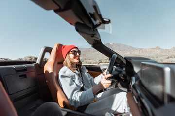 Happy woman in red hat driving convertible car while traveling on the desert road. Carefree...