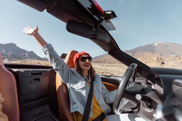 Happy woman in red hat driving convertible car while traveling on the desert road. Carefree lifestyle and travel concept - obrazy, fototapety, plakaty