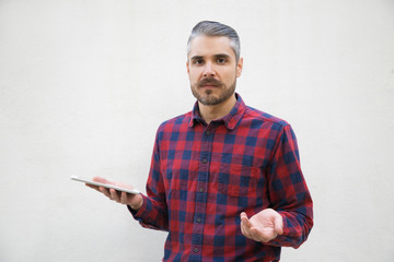 Annoyed young man holding smartphone and looking at camera. Stylish guy having confused look facing...