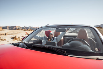 Young woman traveling by convertible car on the picturesquare desert valley, sitting at the passenger seat. Front view through windshield