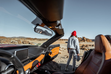 Young woman traveling by convertible sports car, standing with map on the roadside of the desert valley. View from the car interior