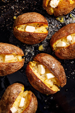 Baked potato, hot baked potatoes with the addition of butter and freshly ground black pepper and sea salt flakes on a black background, top view