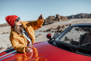 Fototapeta premium Young woman dressed in bright jacket and hat enjoying road trip on the desert valley, standing with phone near the convertible car on the roadside