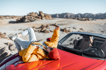 Young woman dressed in bright jacket and hat enjoying road trip on the desert valley, lying on the car hood and photographing on phone