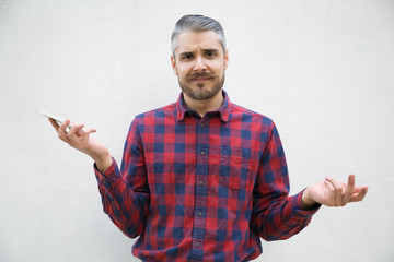 Bearded young man holding smartphone and shrugging shoulders. Stylish guy having confused look facing communication problem while messaging friends through phone. Communication problem concept