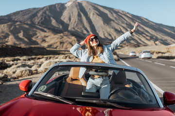 Carefree woman dressed casually in jeans and red hat enjoying road trip on the volcanic valley, raising hands from convertible car on the roadside