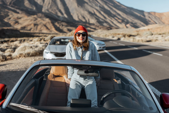 Lifestyle portrait of a young woman enjoying road trip on the desert valley, getting out the convertible car on the roadside