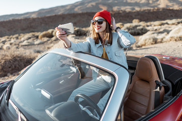Young woman traveling by convertible car on the picturesquare road of the desert valley, photographing or vlogging on mobile phone