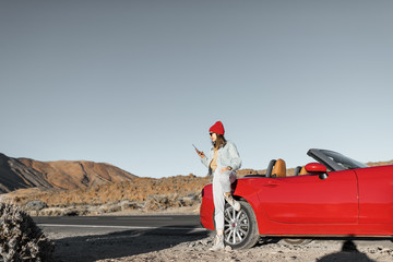 Woman standing with phone on the roadside near the convertible car, traveling on the dessert valley
