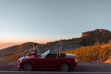 Obraz na płótnie Canvas Landscape view on the roadside above the clouds with woman enjoying beautiful sunset, sitting on the convertible sports car