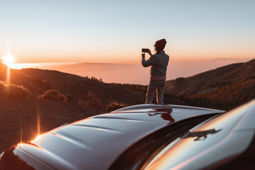 Young woman photographing with phone beautiful landscape during a sunset, sitting on the car hood...