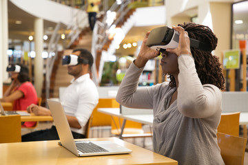Serious African American woman wearing virtual reality glasses.Thoughtful young user sitting in library and experiencing new device. Technology concept