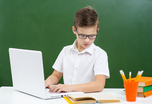 The boy in glasses is engaged in school, sitting on the background of a green blackboard at a laptop and looking at a book