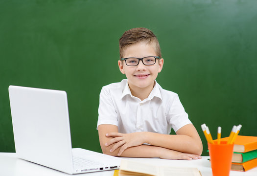 The boy in glasses is engaged in school, sitting on the background of a green blackboard at a laptop