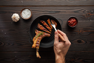 Top view of man holding fork with steak slice by tomato sauce, salt and garlic on wooden background