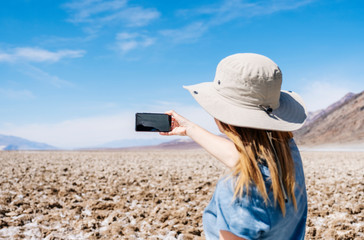 Fototapeta premium A beautiful blond woman with sun hat and yellow and white blue summer dress, takes a selfie photo with the mobile phone in the desert of Death Valley on a sunny day