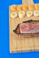 Mediterranean Breakfast. A plate filled with bread and ham, and fruit like tangerine and banana. Blue background.
