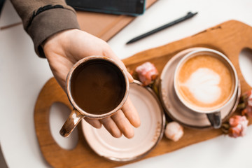 Female hands hold a cup of coffee in their hands over a wooden tray decorated with rosebuds. View from above.