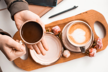 Female hands hold a cup of coffee in their hands over a wooden tray decorated with rosebuds. View from above.