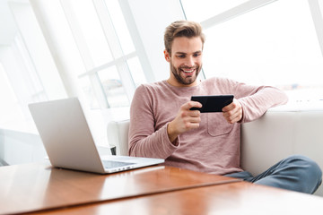 Image of young bearded man using smartphone and laptop in cafe indoors