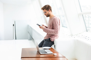 Fototapeta premium Image of young man using smartphone and working on laptop in cafe