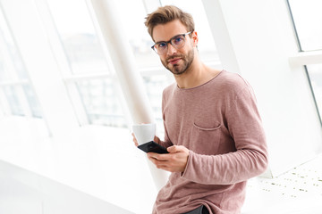 Image of man holding smartphone while standing over window indoors