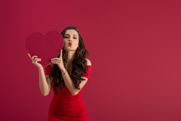 sexy, elegant girl holding paper heart while sending air kiss at camera isolated on red