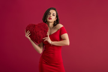 seductive, elegant girl holding decorative heart while looking at camera isolated on red