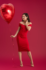 smiling, elegant girl holding heart-shaped balloon while touching neck on red background