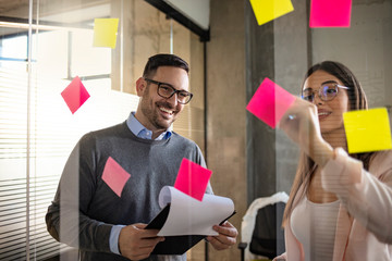 Business people looking at multi colored sticky notes on glass in meeting room at creative office....