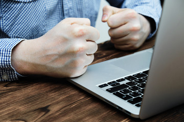 A man sits in a shirt at a laptop clenching his fists. Stress and failure.