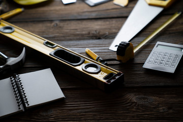 Contractor concept. Tool kit of the contractor: yellow hardhat, libella, hand saw, and notebook on the rustic wooden background.