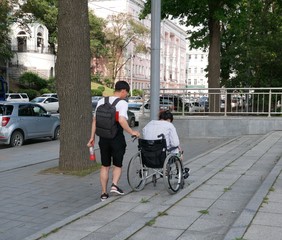 A young man with a black backpack helps a wheelchair user move along the sidewalk of a big city.  Walking around the city on a Sunny summer day.