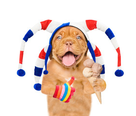 Funny puppy wearing a jester cap holds ice cream and shows thumbs up gesture. isolated on white background