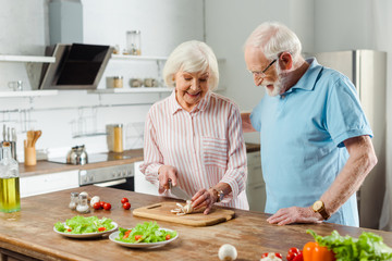 Elderly man hugging smiling wife cutting mushrooms o kitchen table