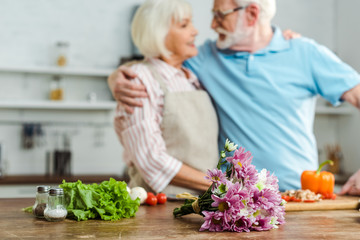 Selective focus of bouquet and fresh vegetables on table and smiling senior couple in kitchen