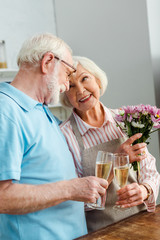 Senior woman with bouquet smiling at husband while clinking with champagne in kitchen