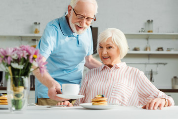 Fototapeta na wymiar Selective focus of senior man putting coffee on table by smiling wife during breakfast