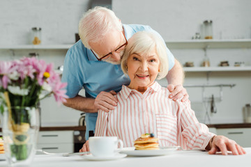 Selective focus of senior man hugging wife looking at camera by pancakes and coffee on table