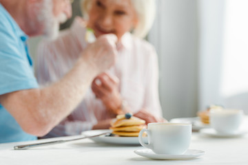 Obraz na płótnie Canvas Selective focus of senior couple smiling at each other by coffee and pancakes on table