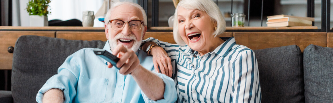 Panoramic shot of senior couple laughing while watching tv on sofa at home