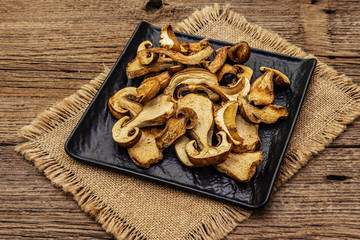 Fototapeta na wymiar Dried porcini mushrooms. Traditional ingredient for cooking healthy food. Black ceramic plate, sackcloth napkin, old wooden boards background