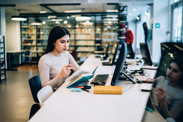 Thoughtful female student reading textbook and using computer while studying in library