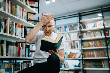 Amazed crouched student holding opened book and touching forehead in library