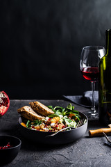 Buckwheat salad with lamb's lettuce, pomegranat seeds, goat cheese, mandarine and spring onion, Served with whole grain baguette and red wine. Black table and black background.