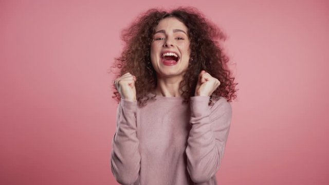 Girl with curly hair and extraordinary makeup shows yes gesture of victory, she achieved result, goals. Surprised excited happy woman on pink background