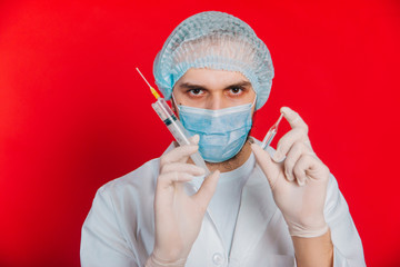 The doctor is holding a syringe and an ampoule with a vaccine. A young guy in a white coat, in a medical mask and gloves on a red background. Medical worker in a cap.
