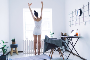 Relaxed woman stretching up while standing by window in morning